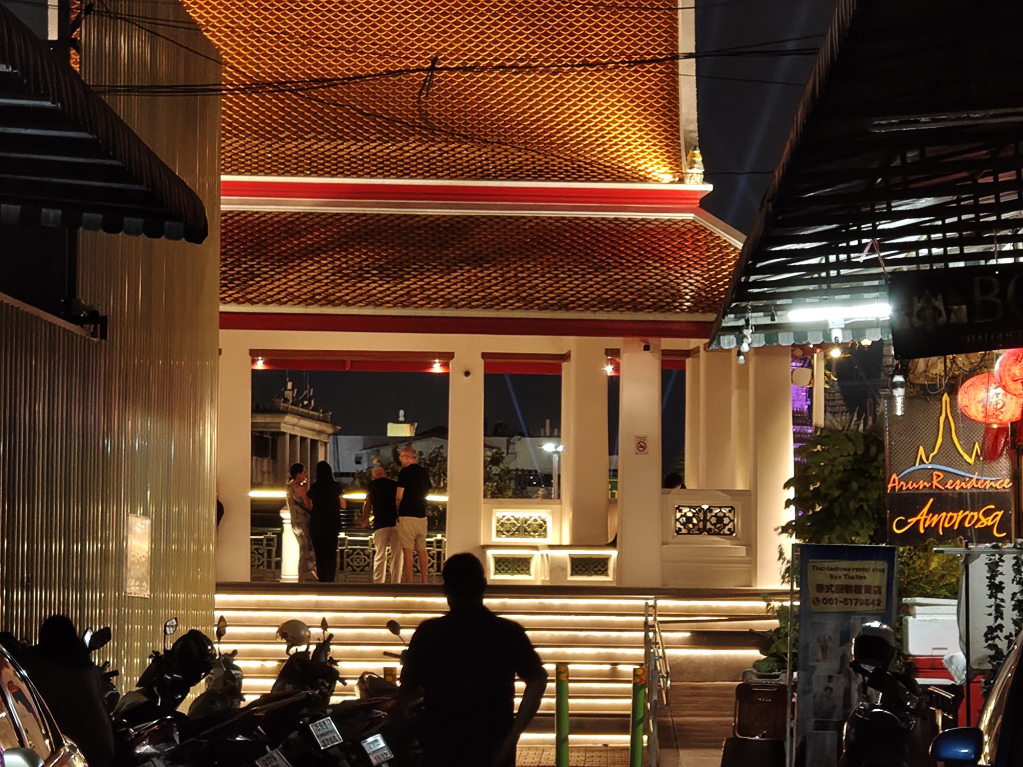 A gilded temple roof at night with motorbikes parked in front and someone walking past